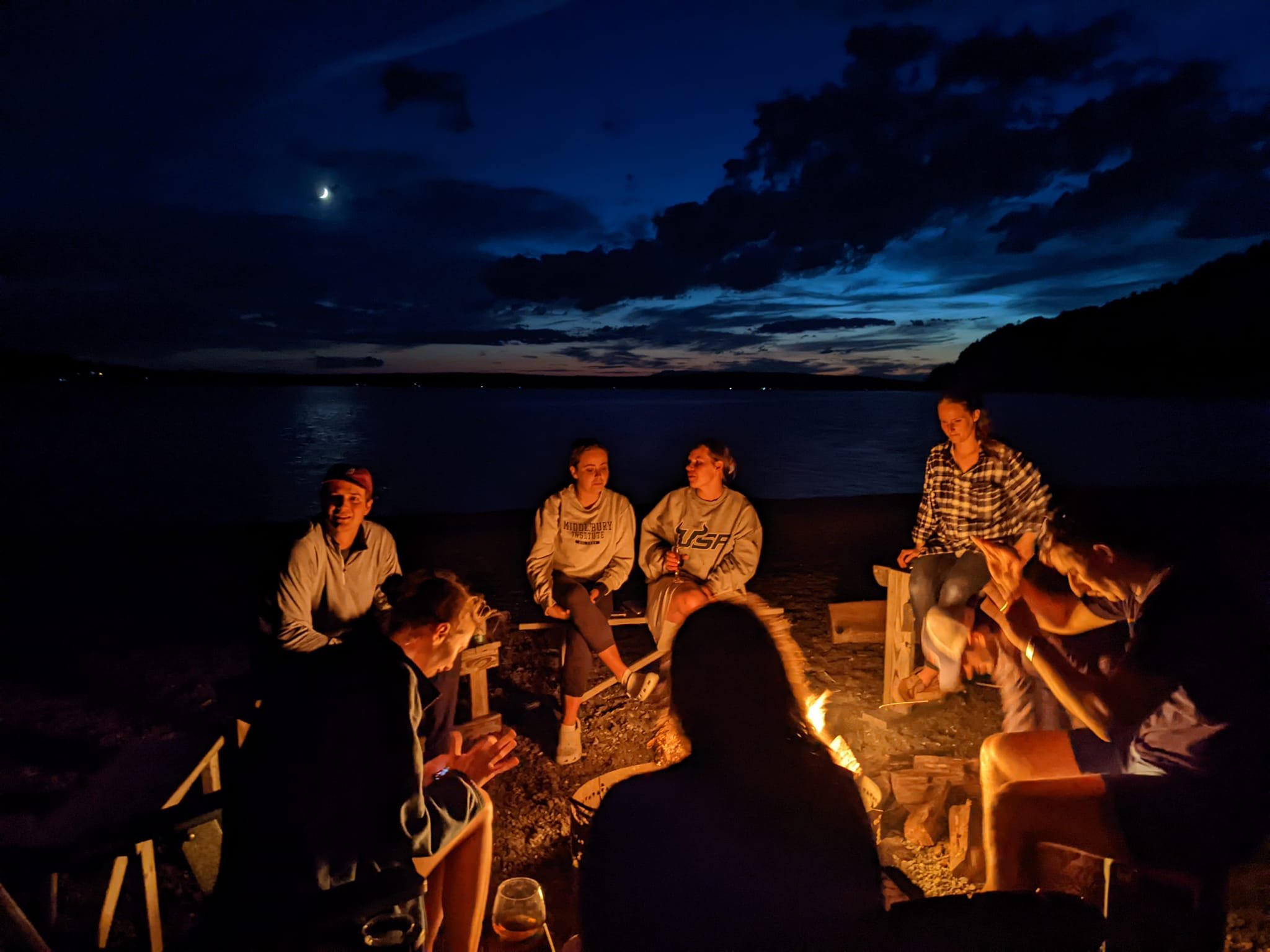 Group of people sitting around a campfire at dusk on a beach, with a dark sky, crescent moon, and a body of water in the background.