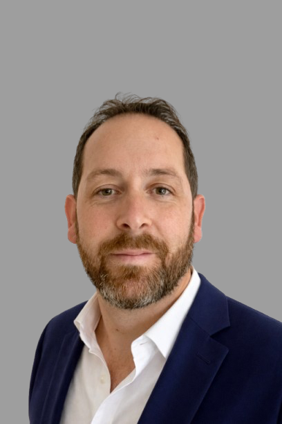 A man with short, dark, and slightly curly hair wearing a blue and white gingham button-up shirt, posing with a neutral expression against a gray background.