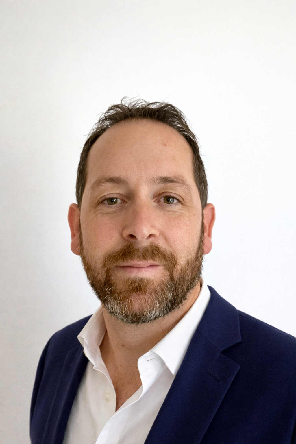 A man with short, dark, and slightly curly hair wearing a blue and white gingham button-up shirt, posing with a neutral expression against a gray background.