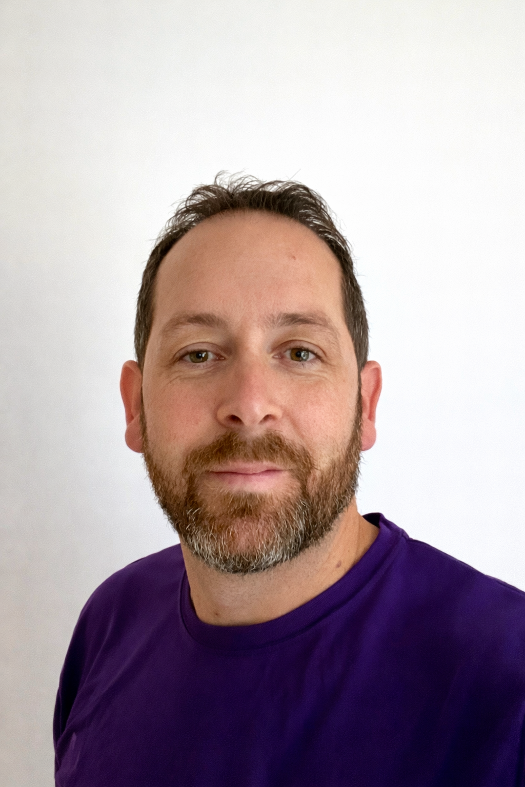 A man wearing a purple shirt with a logo featuring a unicorn and text that says 'VMS' on the front, standing against a gray background.
