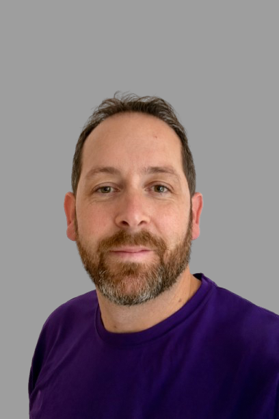 A man wearing a purple shirt with a logo featuring a unicorn and text that says 'VMS' on the front, standing against a gray background.