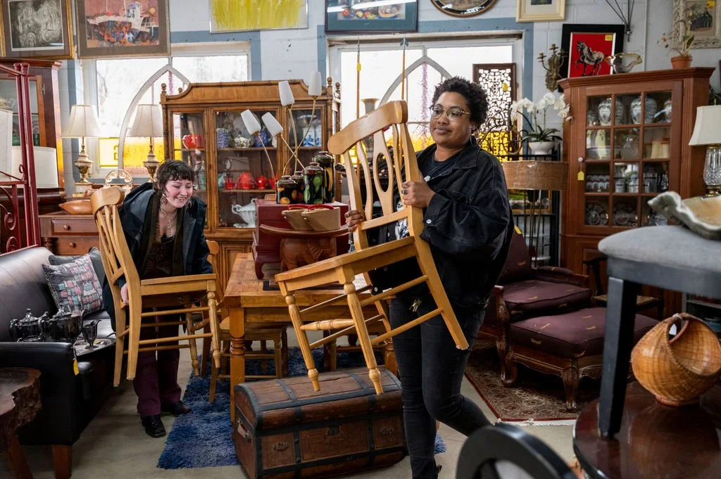 Two women are in an antique store, each holding a wooden chair. The woman on the right, with curly hair and glasses, wears a black jacket. The woman on the left, with short dark hair, is smiling and leaning forward. The store is filled with furniture, lamps, and decorative items.