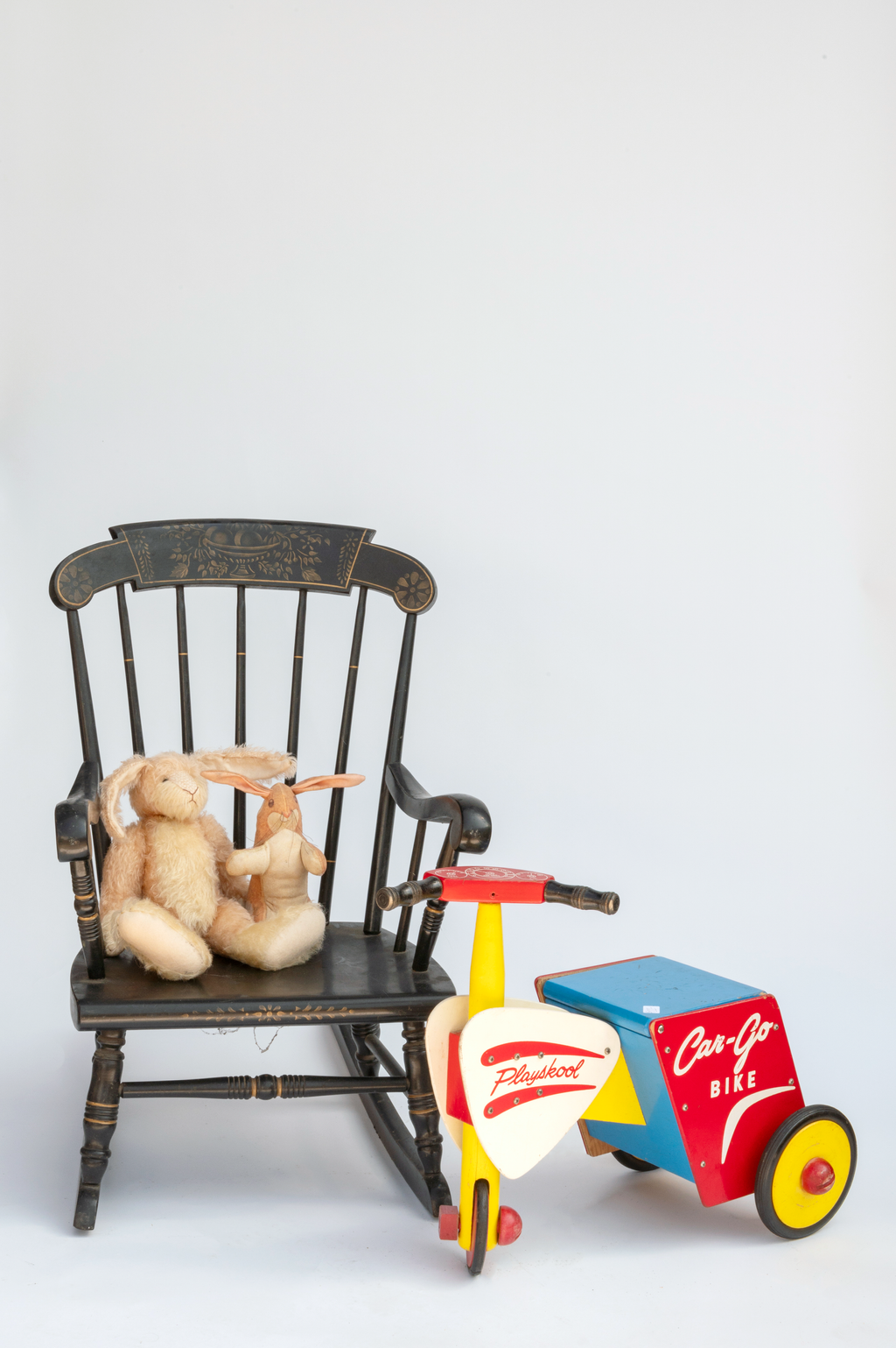 Stuffed toys, a plush bunny and a rabbit, seated on a vintage wooden rocking chair with floral design, next to a colorful wooden toy bike with a sidecar labeled "Car-Go Bike" and "Playkool".