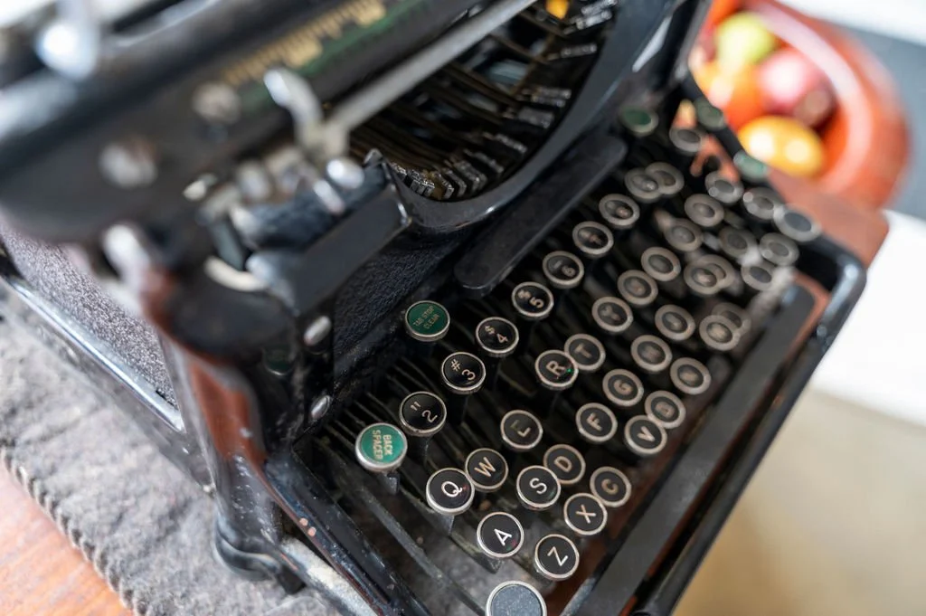 Close-up of an antique black typewriter with round keys, placed on a gray textured surface near a bowl of colorful fruit.