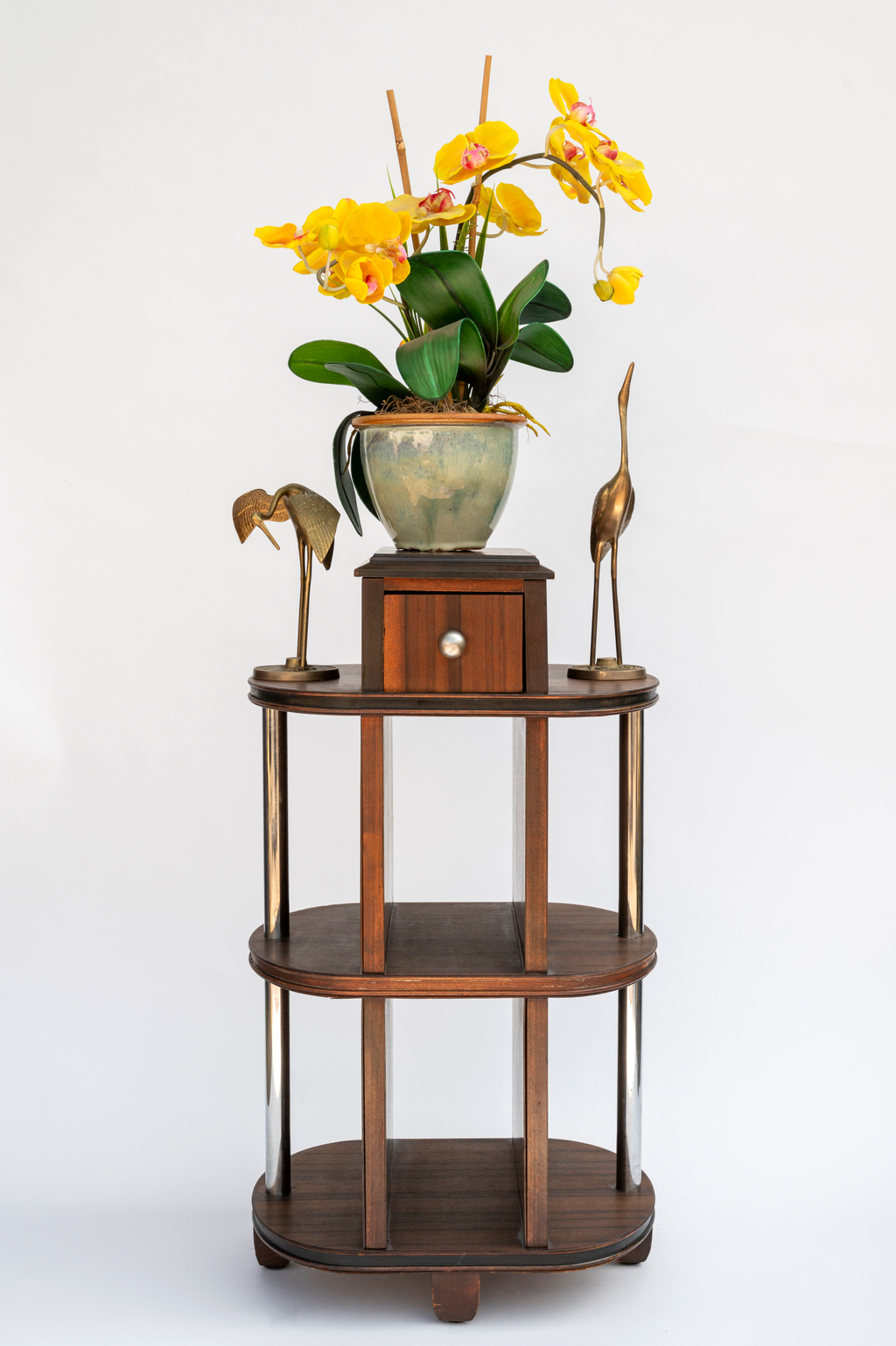 Wooden tiered plant stand with a potted yellow orchid and decorative brass bird sculptures against a white background.