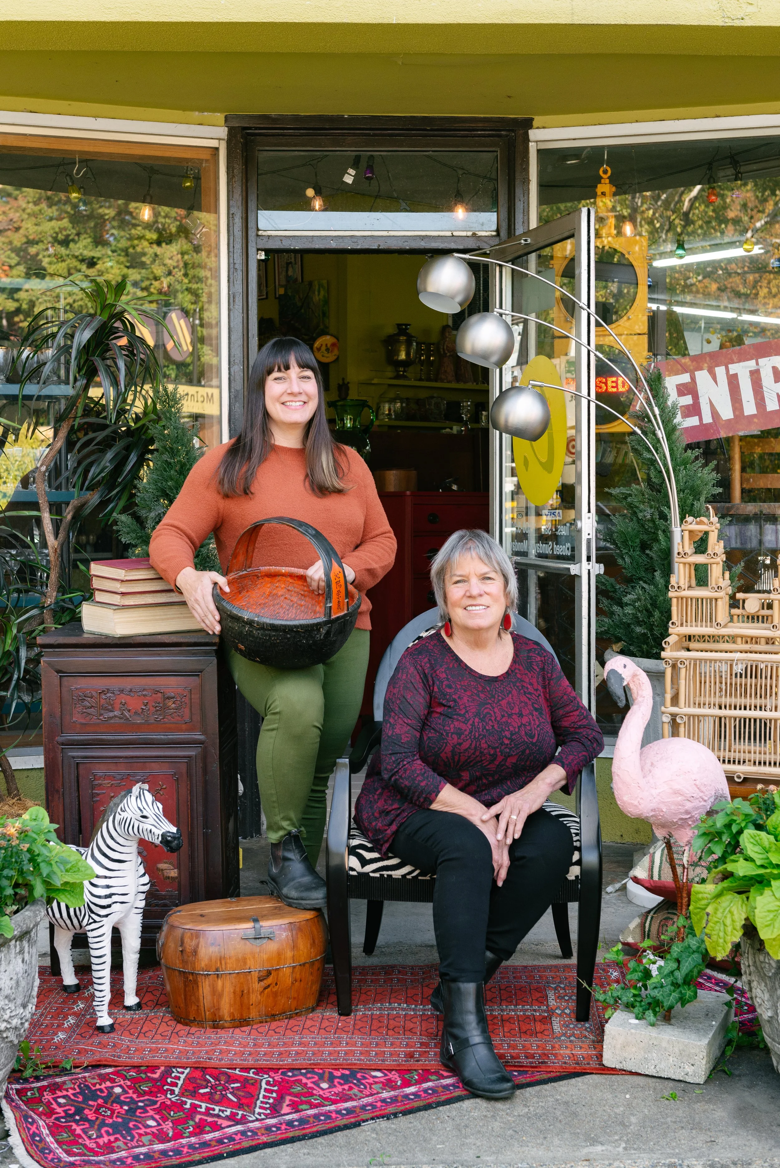 Circa owners posing in front of the colorful antique store entrance, surrounded by decorative items including a zebra, a flamingo, books, and plants.