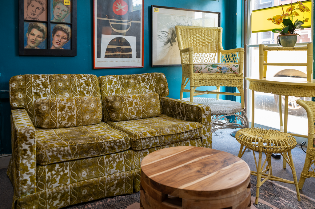 A vintage-style interior with a patterned green and yellow sofa, rattan chairs, a wooden coffee table, painting art on teal walls, and a potted orchid near a window with yellow blinds.