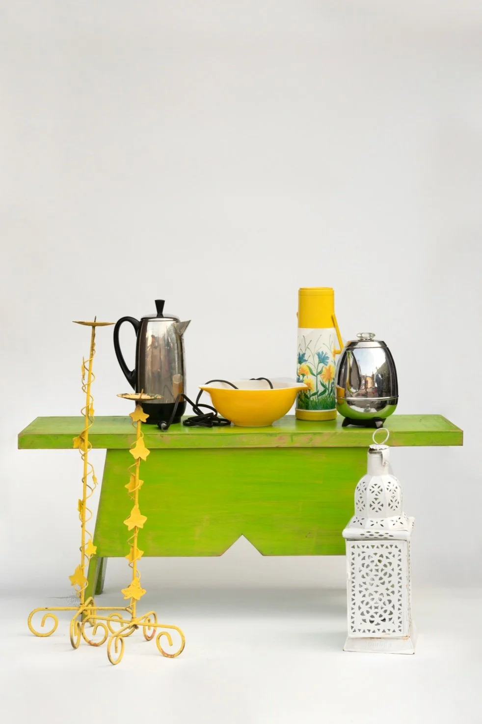 Green wooden table with kitchen appliances and decorative items, including a yellow bowl, electric kettle, yellow thermos, metallic toaster, white lantern, and yellow stand with star-shaped hanging ornaments, all against a plain white background.