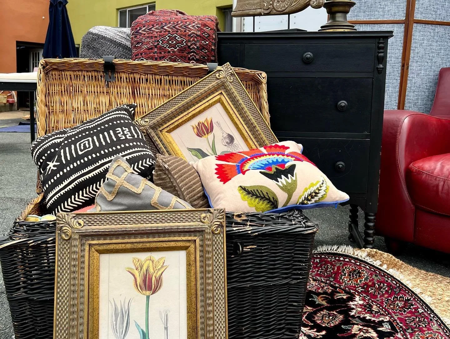 A wicker basket filled with decorative pillows, art prints, a black dresser, and a red leather armchair in an outdoor setting.