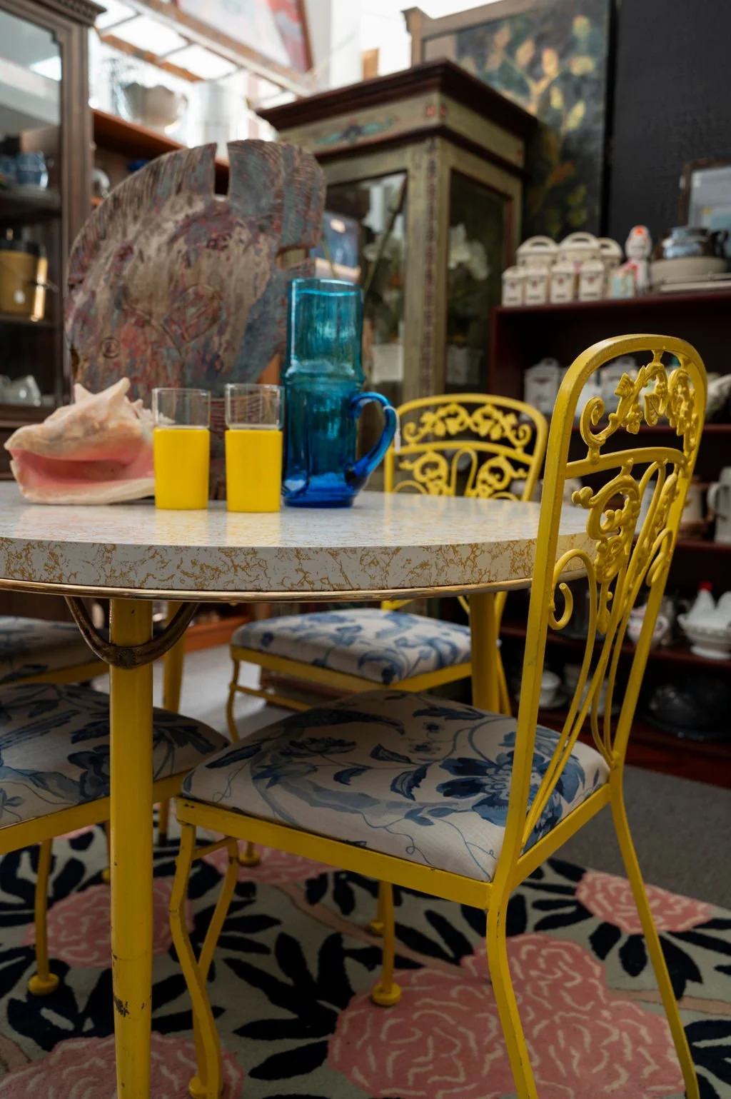 Colorful vintage dining space with a yellow metal chair with floral cushion, a white marble table, and various decorative items including blue and yellow glassware, a seashell, and an abstract art piece. Background features shelves with china and a distressed cabinet.