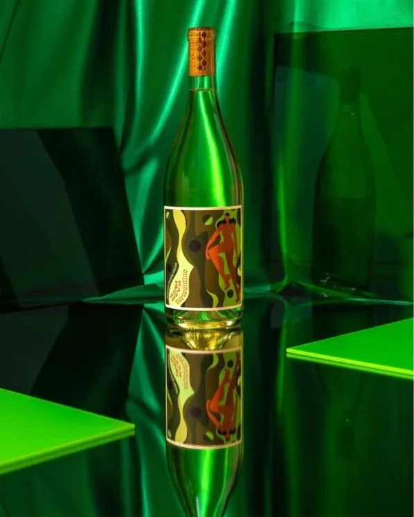 A green wine bottle with a colorful abstract label, featuring red and green patterns, on a reflective surface with green drapery and geometric shapes in the background.