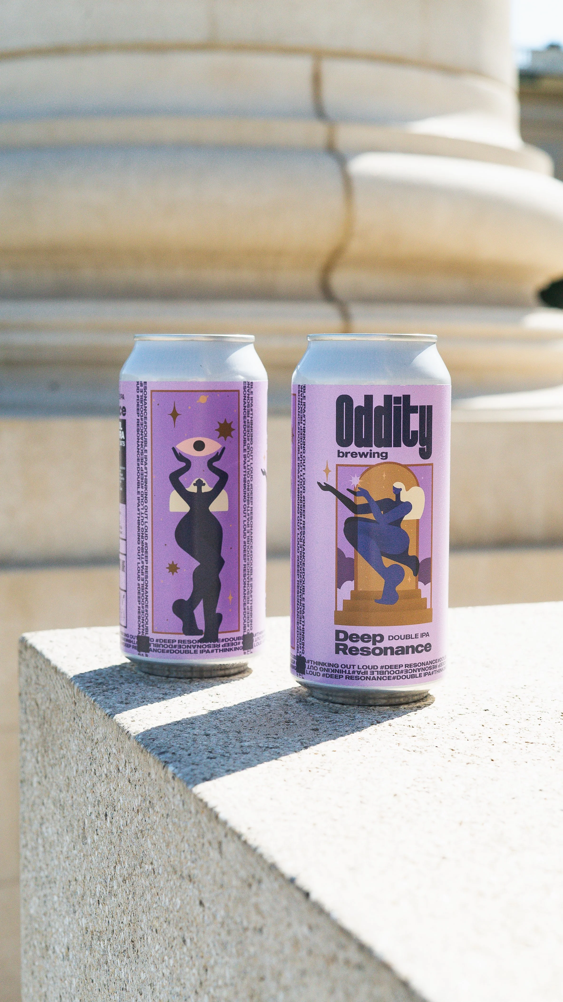 Two cans of Oddity Brewing's "Deep Resonance" Double IPA, featuring artistic labels with abstract figures and celestial elements, placed on a concrete surface in outdoor lighting.