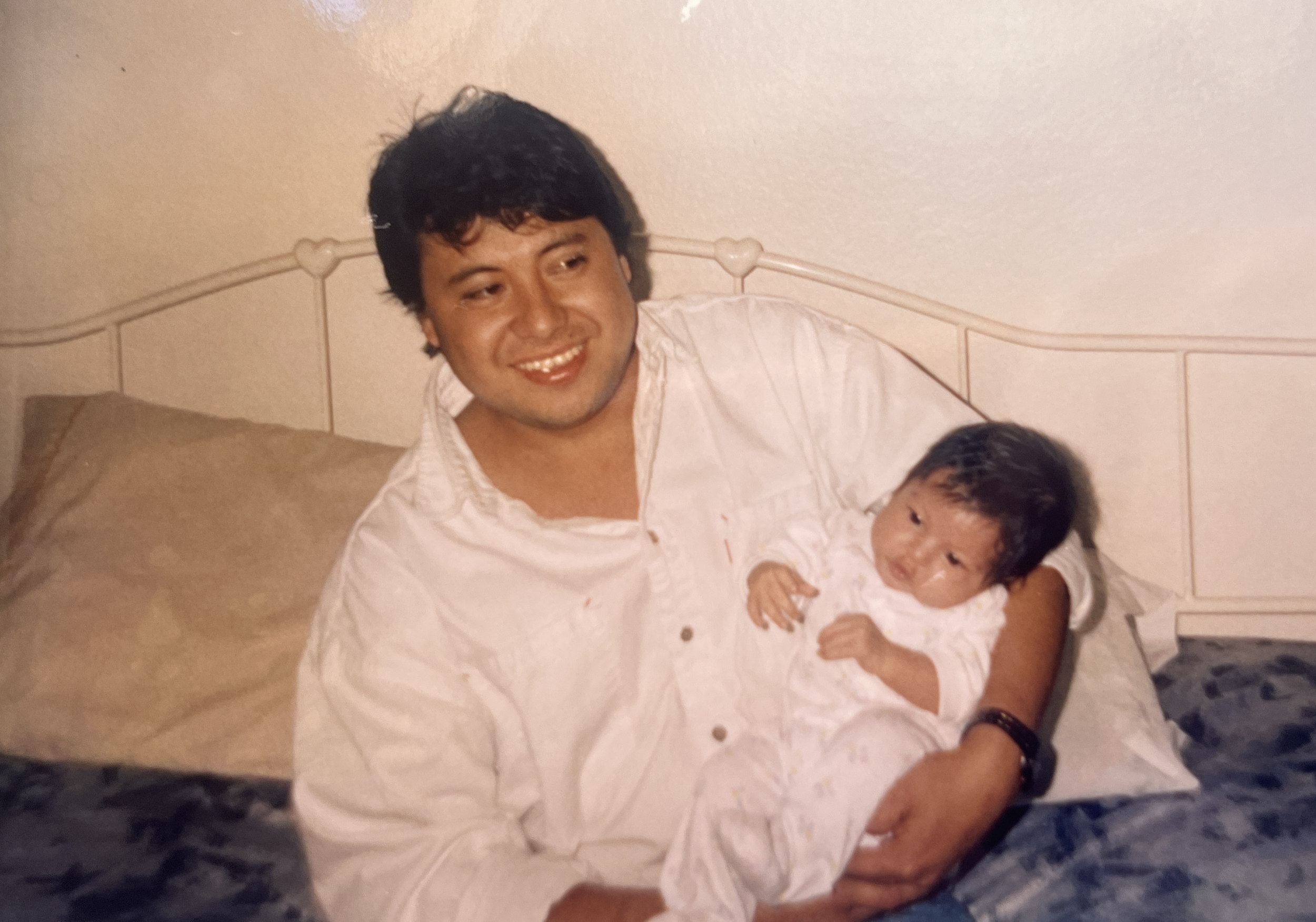 An adult man with dark, curly hair and a smile, wearing a white shirt, holds a baby with dark hair, lying on a bed with beige pillows and a cream-colored headboard in the background.