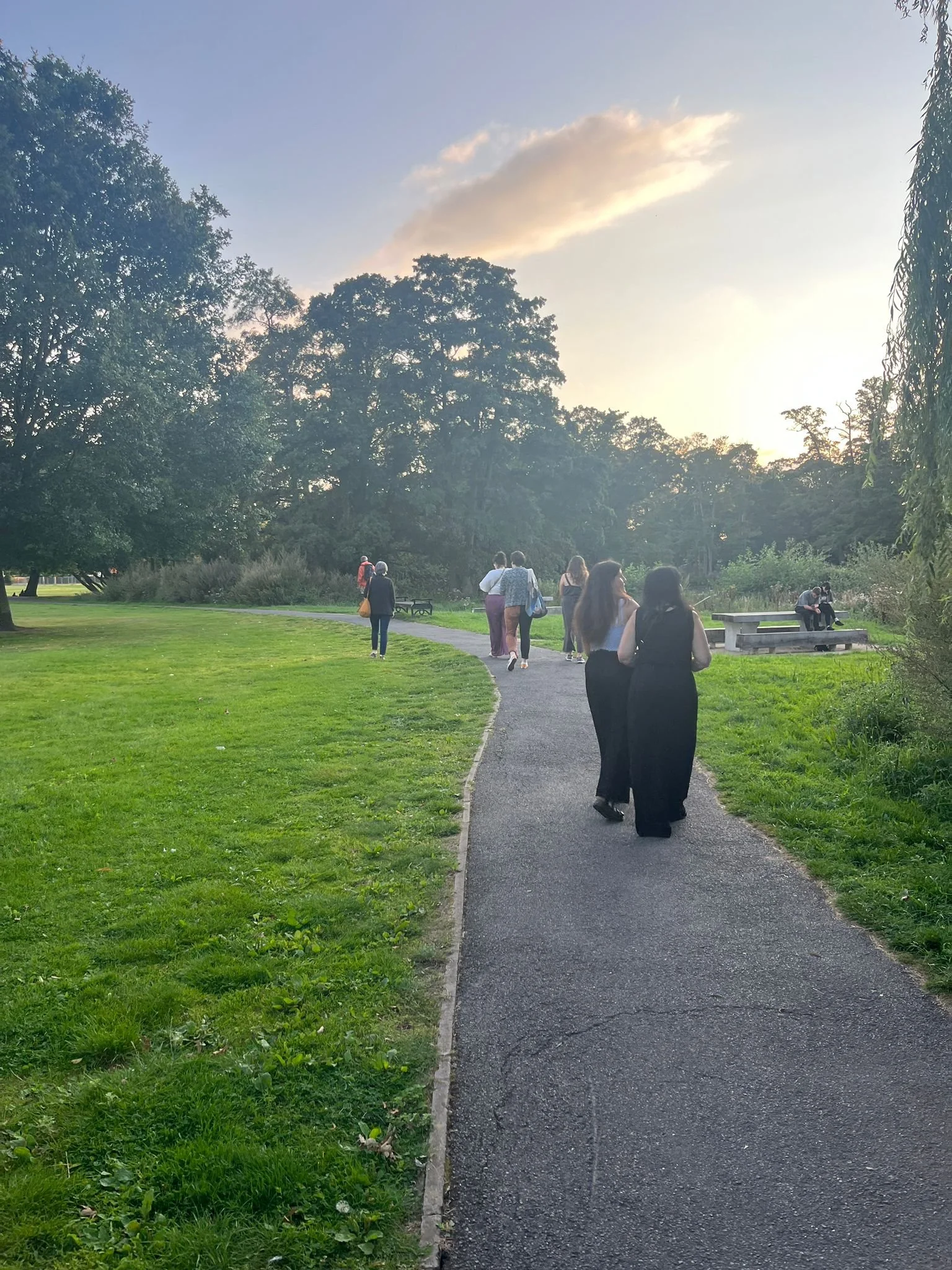 People walking on a paved path at boultham park during sunset, with green grass and trees surrounding them.