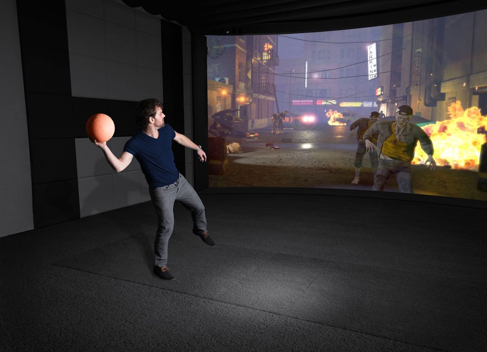 A man in a dark blue shirt and gray pants stands inside a virtual reality room, holding a basketball in his right hand, with a large screen displaying a chaotic street scene with explosions and zombies in front of him.