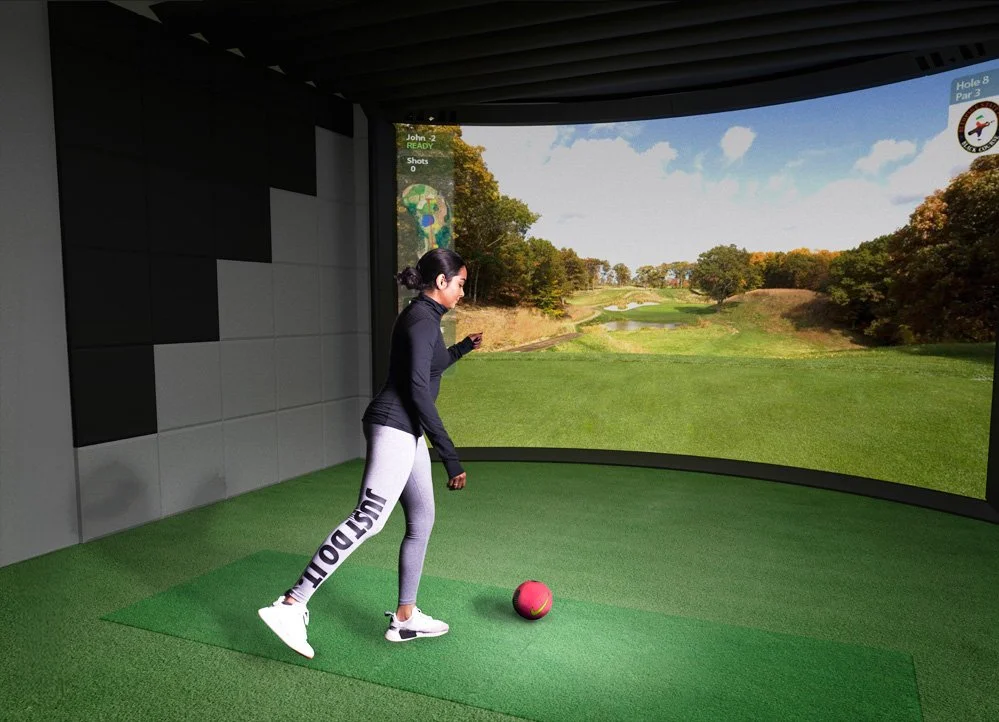 Woman standing on artificial green grass in an indoor golf simulator, preparing to hit a pink golf ball. The simulator displays a scenic golf course with trees and a blue sky on the screen behind her.