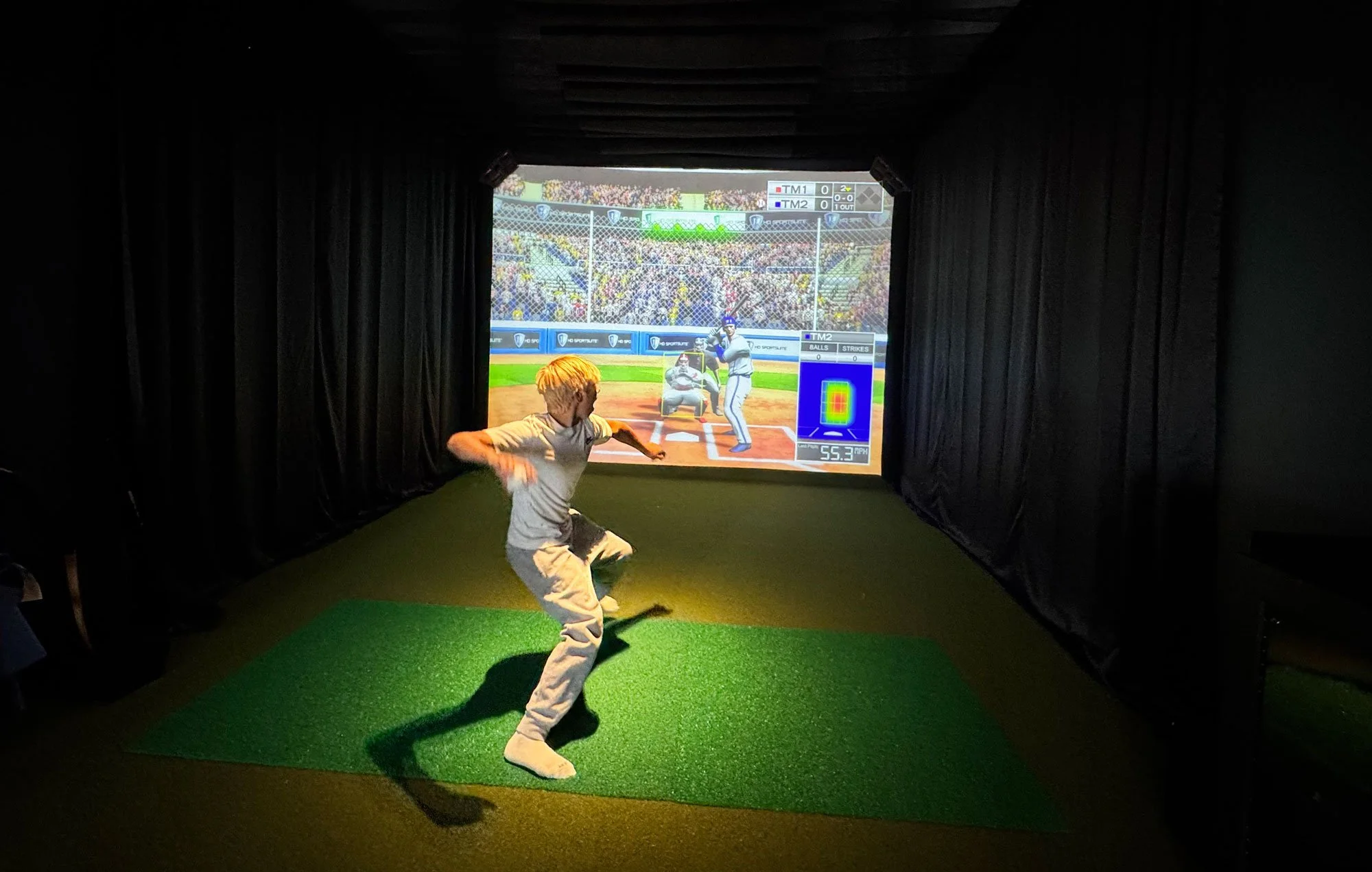 A boy practicing baseball swings in an indoor batting cage with a large screen displaying a baseball game projection behind him.