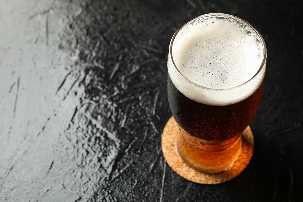 A tall glass of dark beer with a frothy head on top, placed on a cork coaster on a black textured surface.