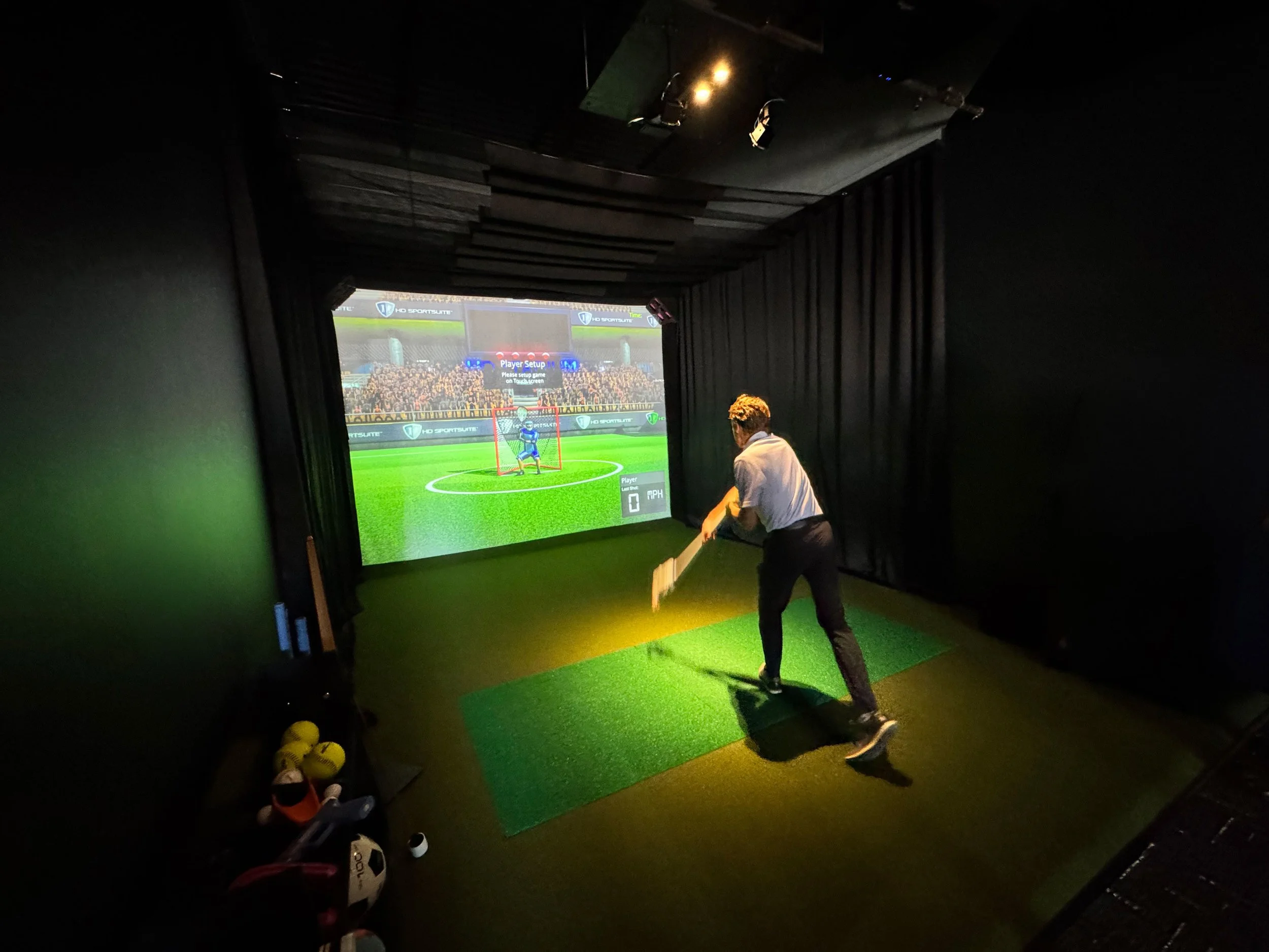 An indoor golf simulation room with a large screen showing a virtual soccer field and a player setup interface, a person preparing to hit a golf ball, and scattered golf balls and shoes on the floor.