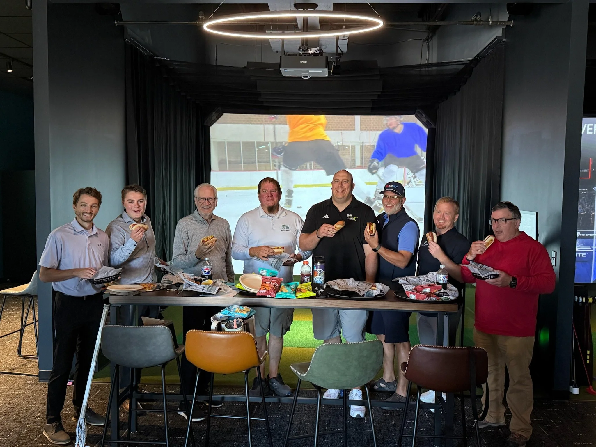 Group of men holding hot dogs inside a room with black curtains, with an ice hockey game playing on a large screen in the background.