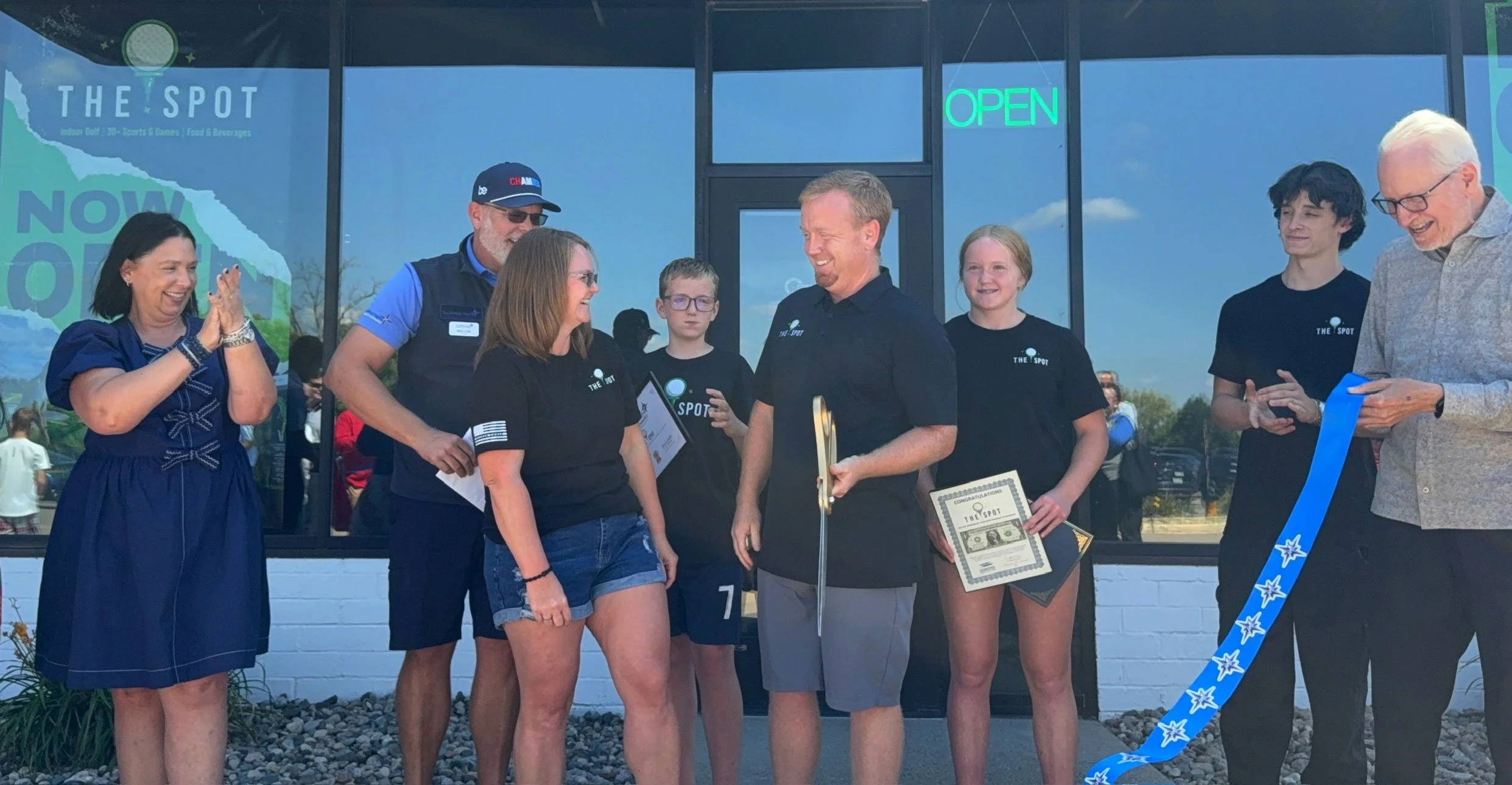 People celebrating the opening of a new business called 'The Spot' with a ribbon-cutting ceremony outside the storefront, with a woman in a blue dress clapping and others smiling and holding certificates and scissors.