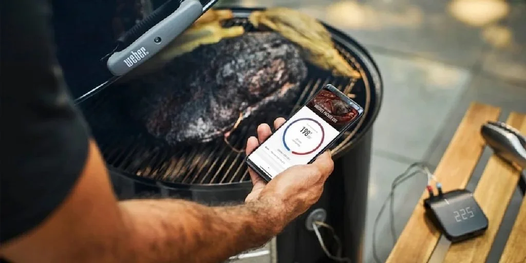 Person using a smartphone to monitor the temperature of a steak on a Weber grill.
