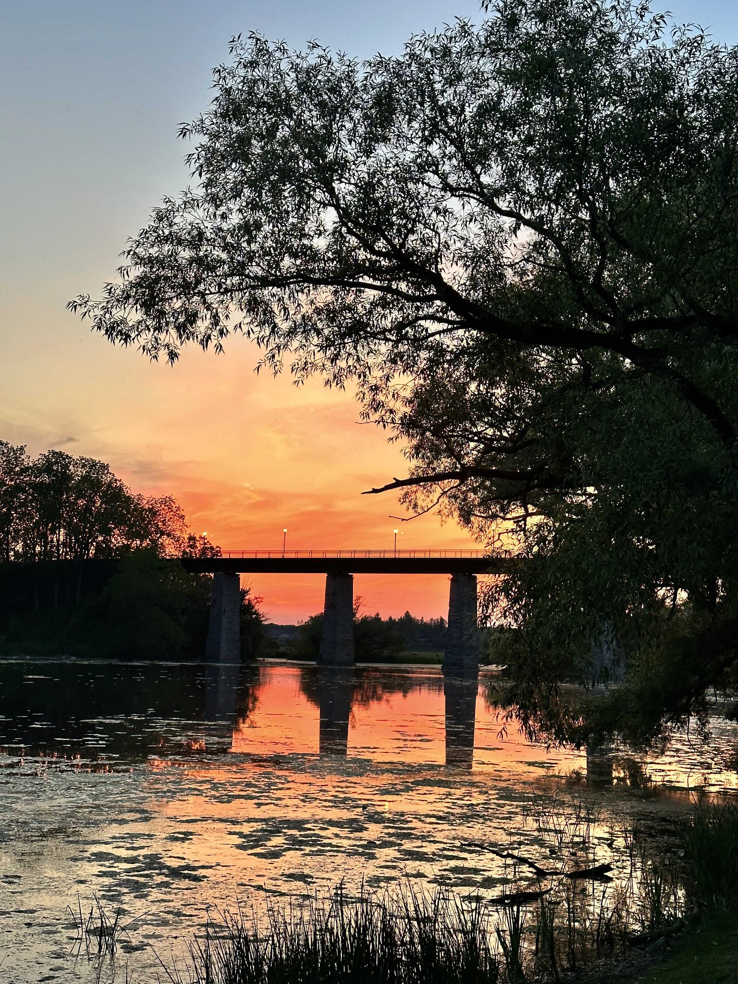 A river at sunset with a bridge in the background and trees along the shore. The sky is orange and pink, reflected in the water.
