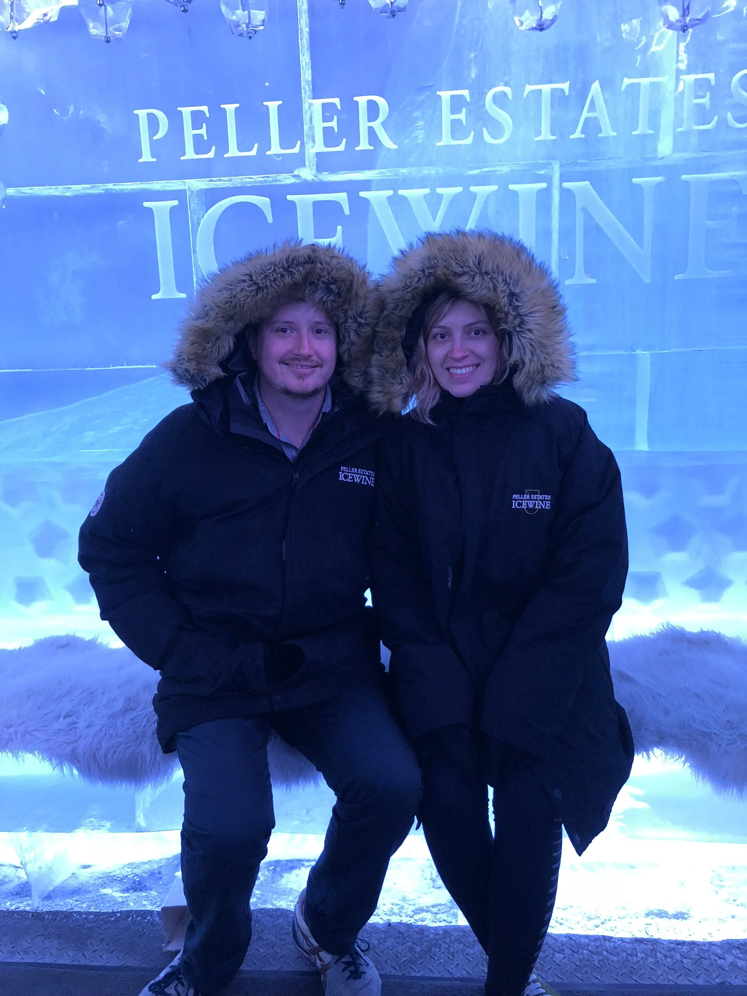 A man and woman dressed in black winter jackets with fur-lined hoods sitting on a bench inside an ice-themed exhibit at Peller Estates Icewine. The background features large ice blocks with the words "Peller Estates Icewine" engraved on them.