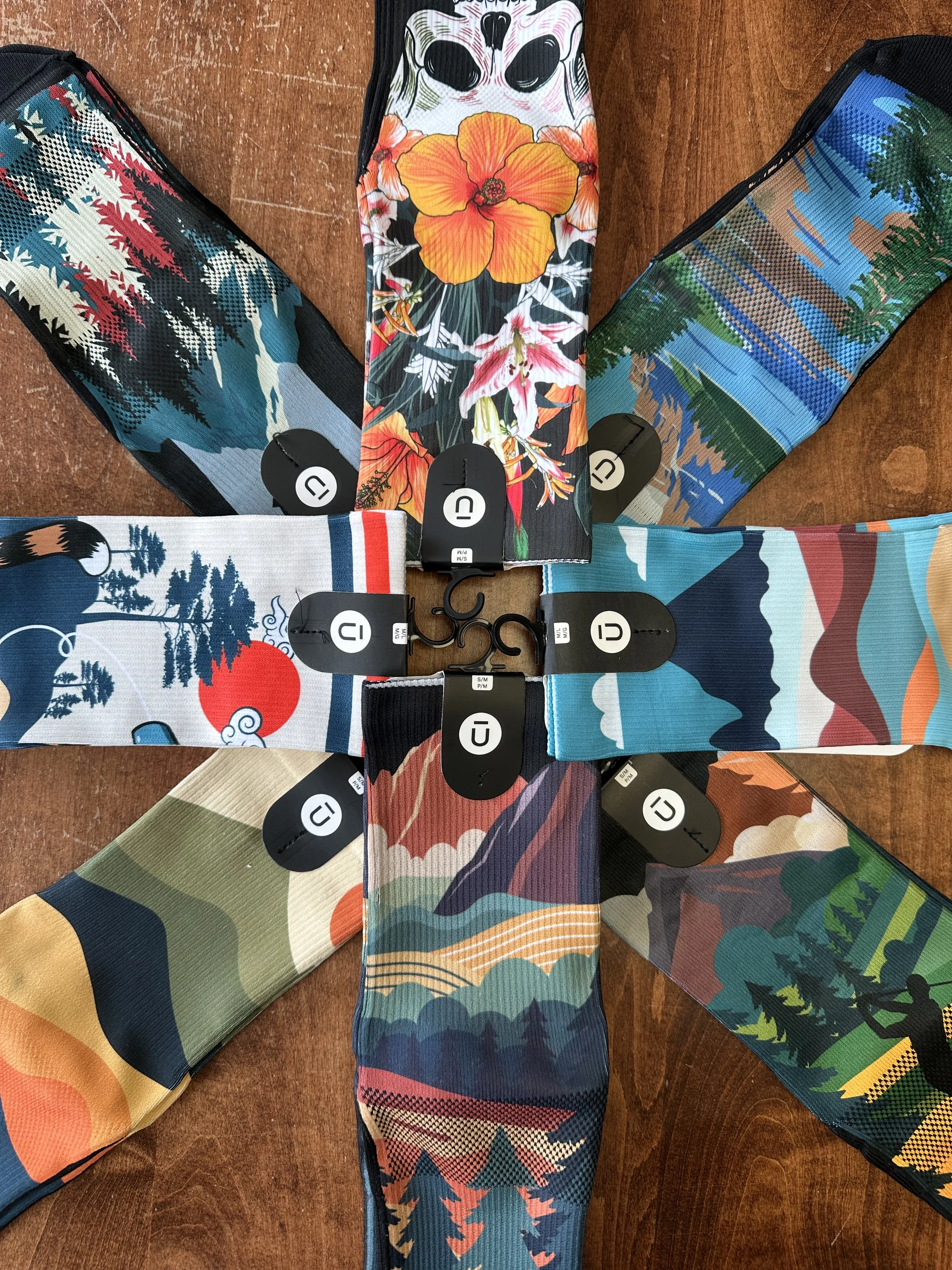 A collection of colorful patterned fabric socks arranged in a circular pattern on a wooden surface, with tags attached to each sock.
