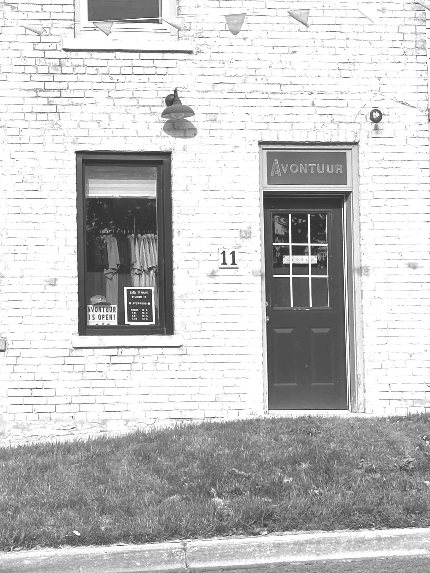 Black-and-white photo of the exterior of a brick building with a door and a window. The window displays clothing and signs. The door has the word 'AVONTUUR' above it, and there is a street number '11' on the wall.