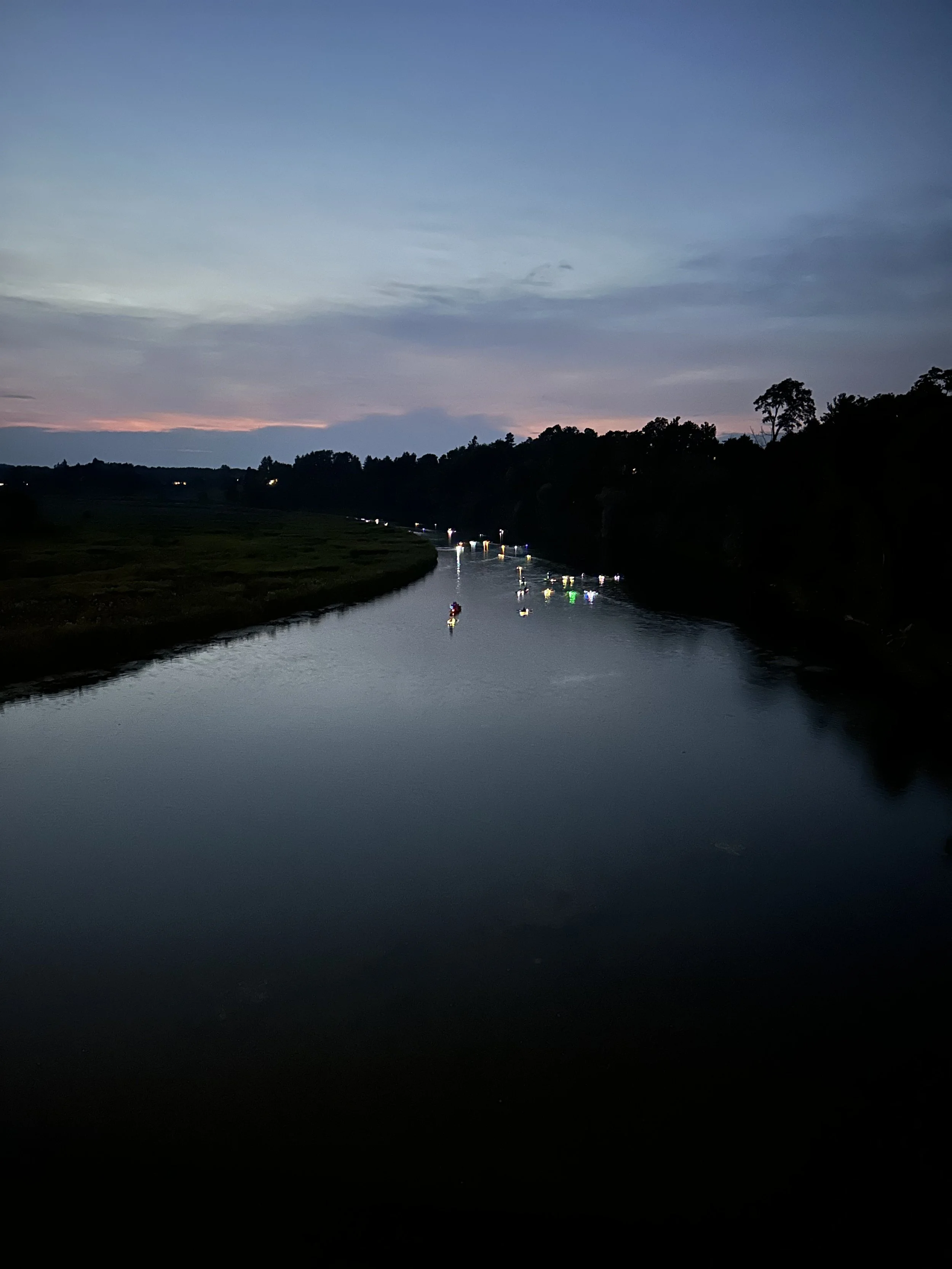 A river at dusk with boats and lights on the water, trees on the riverbanks, and a partially cloudy sky with a faint pinkish hue near the horizon.