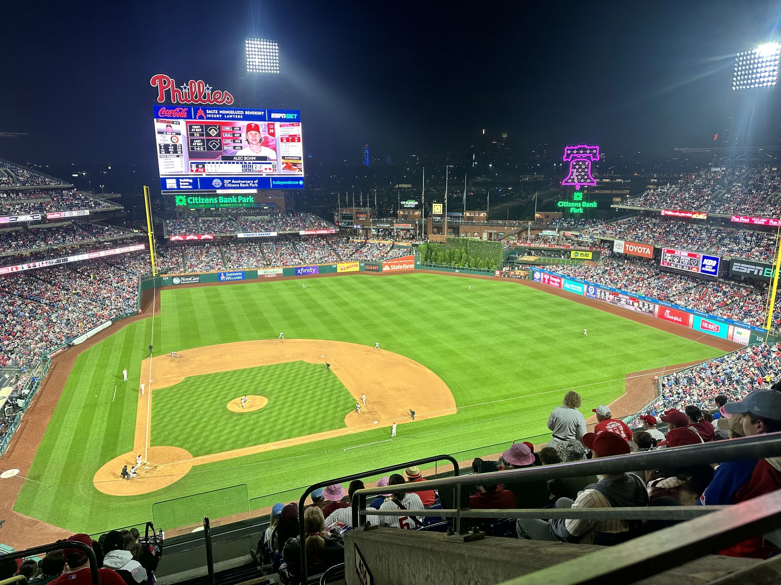 Nighttime view of a baseball stadium filled with spectators, with players on the field, advertising billboards, and a large scoreboard displaying player information, with a large crowd and bright stadium lights.