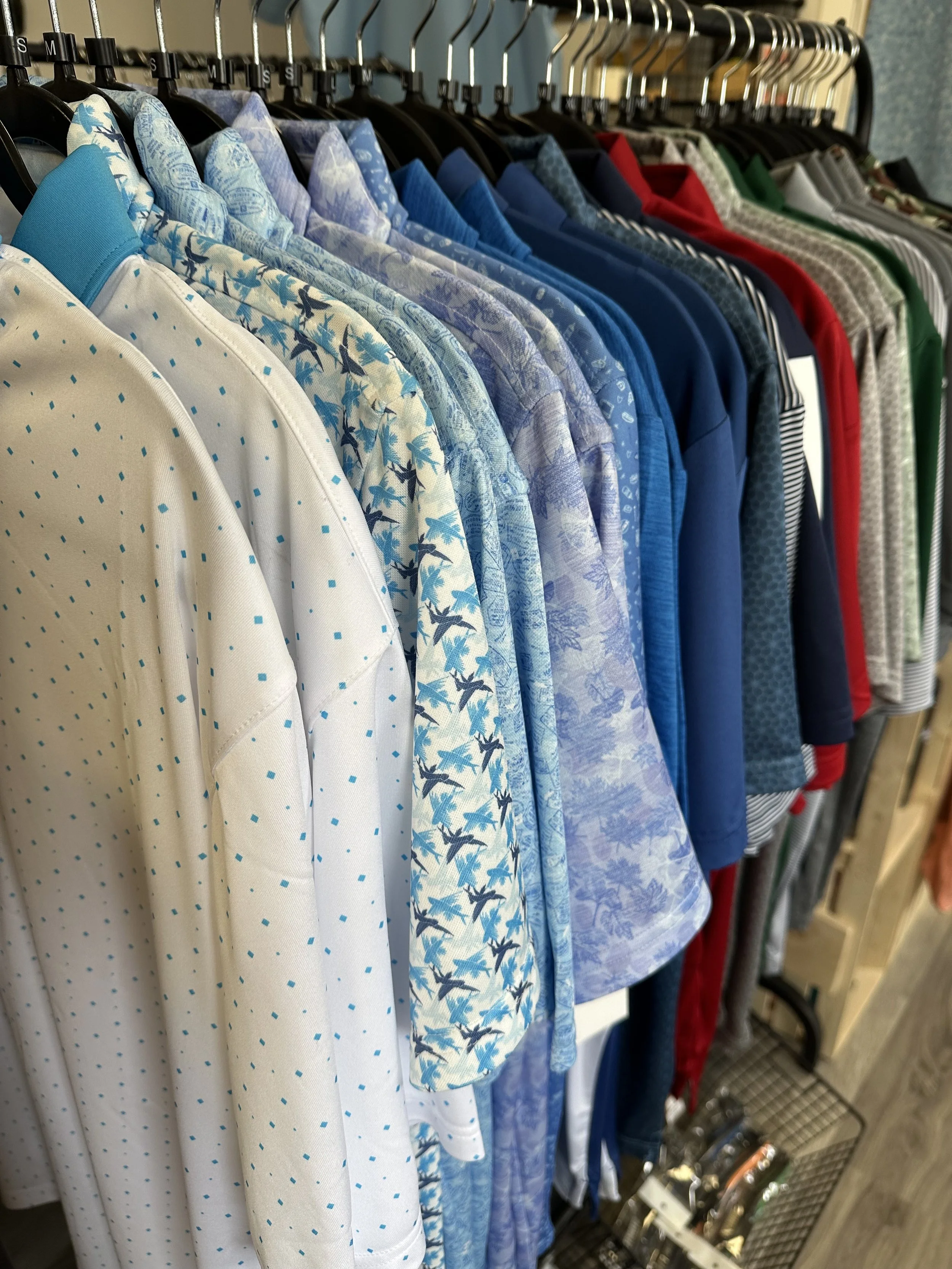 A rack of men's polo shirts in various colors and patterns, including white with blue dots, blue palm trees, shades of blue, red, green, and gray, hanging on black hangers.