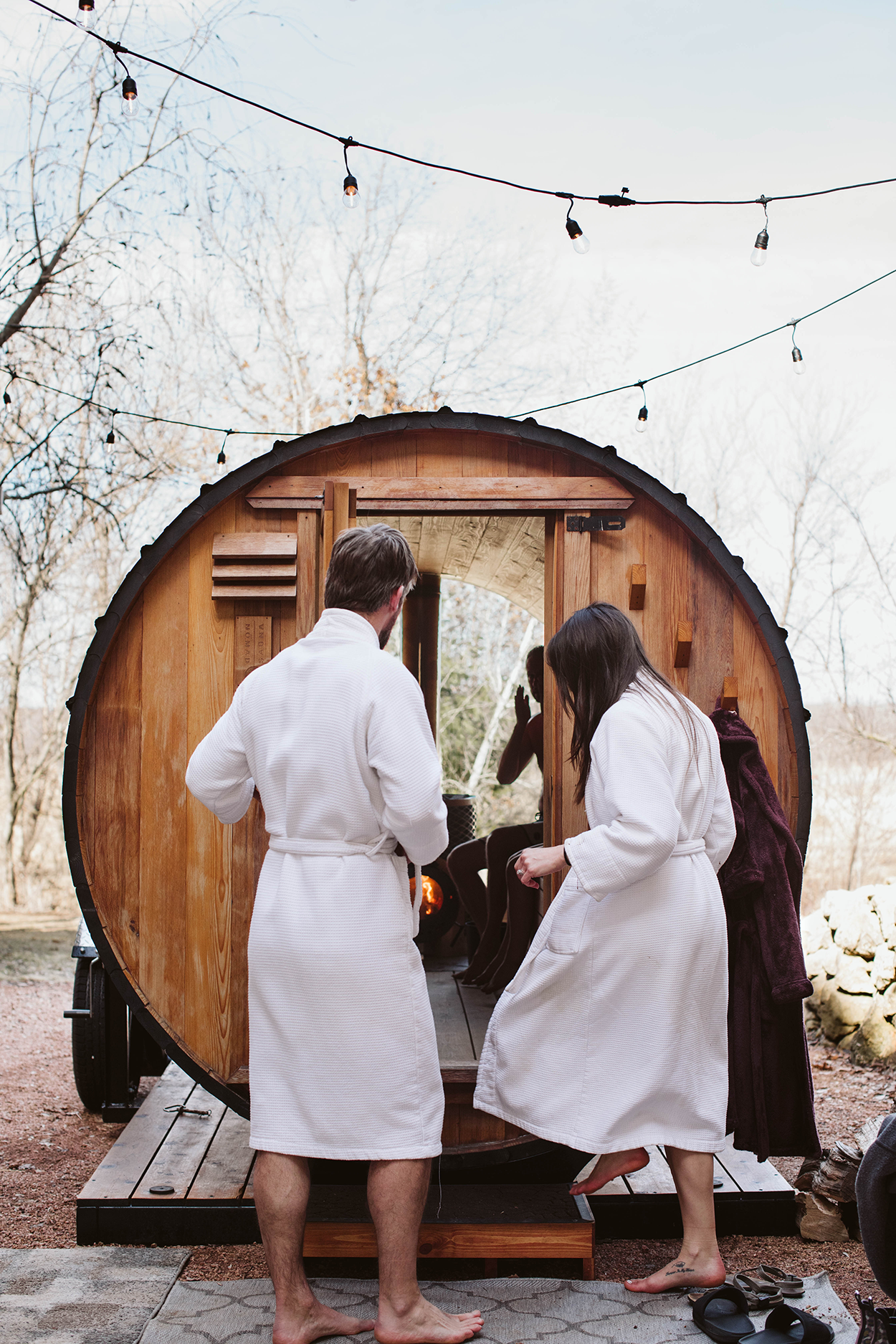 Man and woman about to enter a wood fired sauna in white robes.