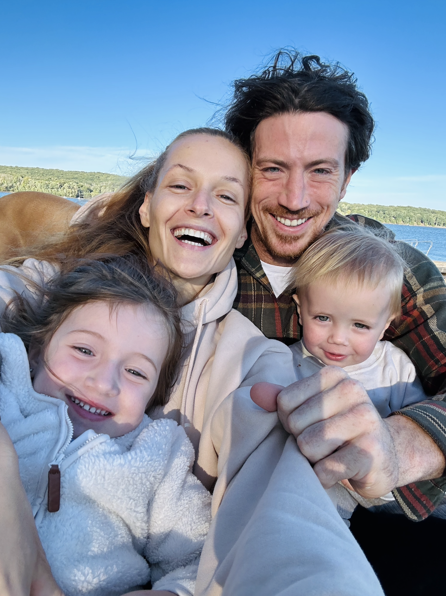A happy Foley family, including their two young children, taking a selfie outdoors near a lake, with trees and blue sky in the background.