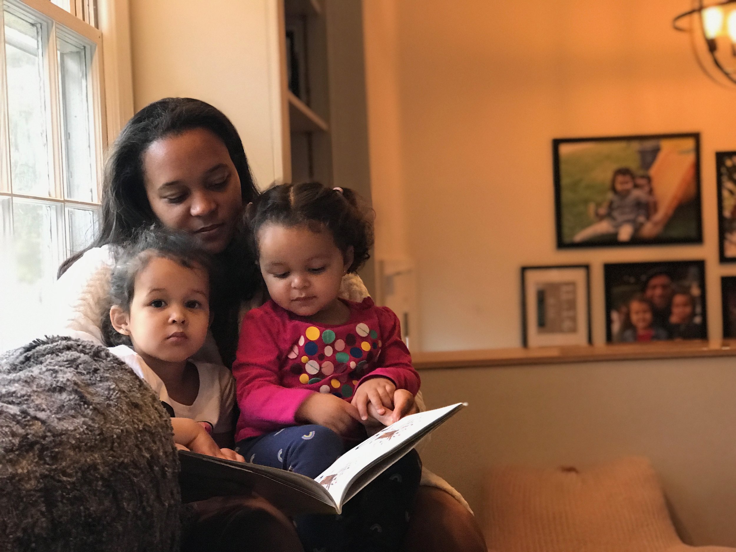 A woman with dark hair and a white sweater sitting with three young children in a cozy home setting, looking at a photo album or book together.