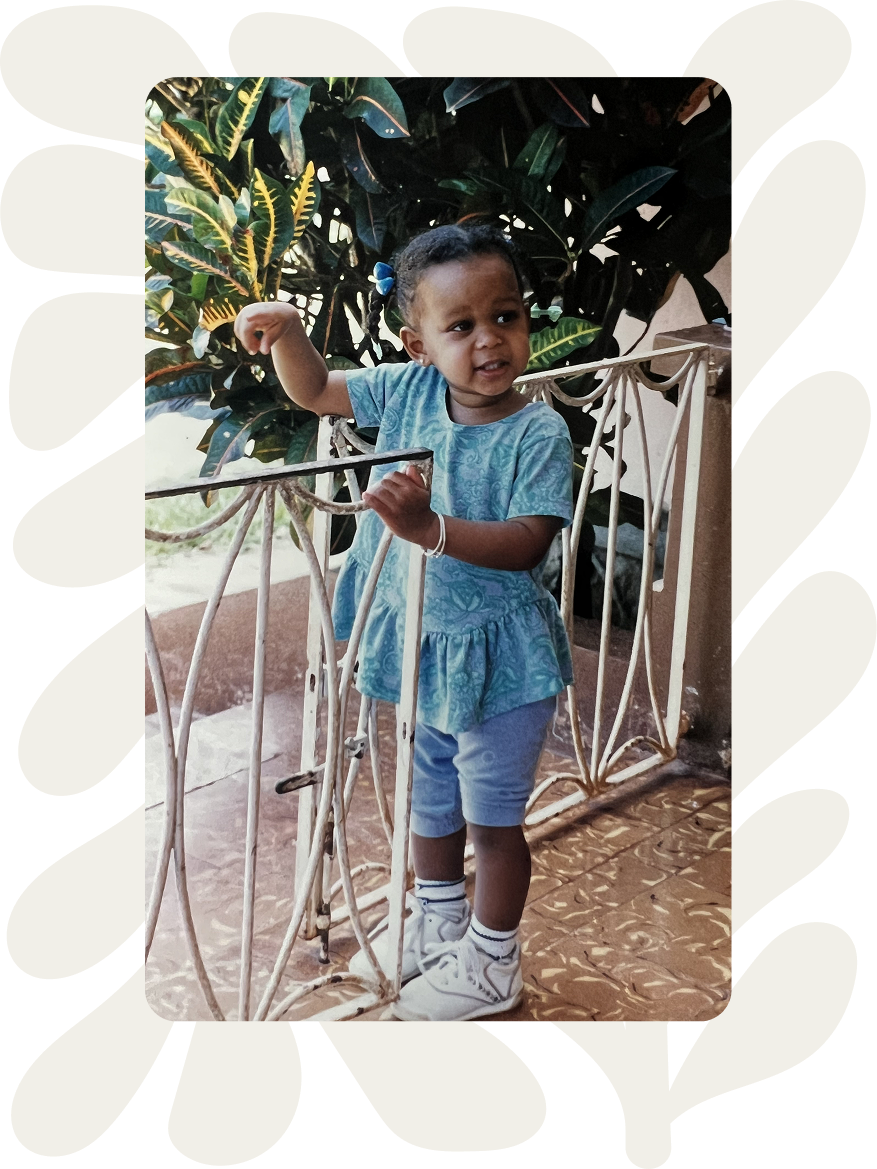 A young girl standing behind a white metal gate outdoors, with large green leafy plants in the background. She is wearing a light blue dress, white socks, and white sneakers, and has a blue bow in her hair. She is holding onto the gate with one hand and appears to be making a facial expression of confusion or concern.