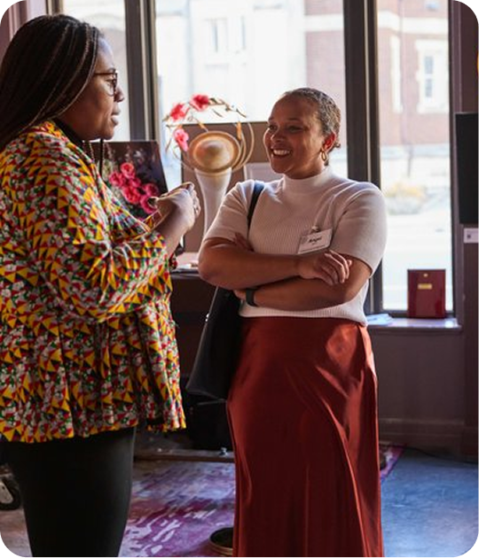 Two women are having a conversation in a brightly lit room near a window. One woman is wearing glasses and a colorful patterned jacket, while the other is smiling and wearing a white top and a long red skirt.