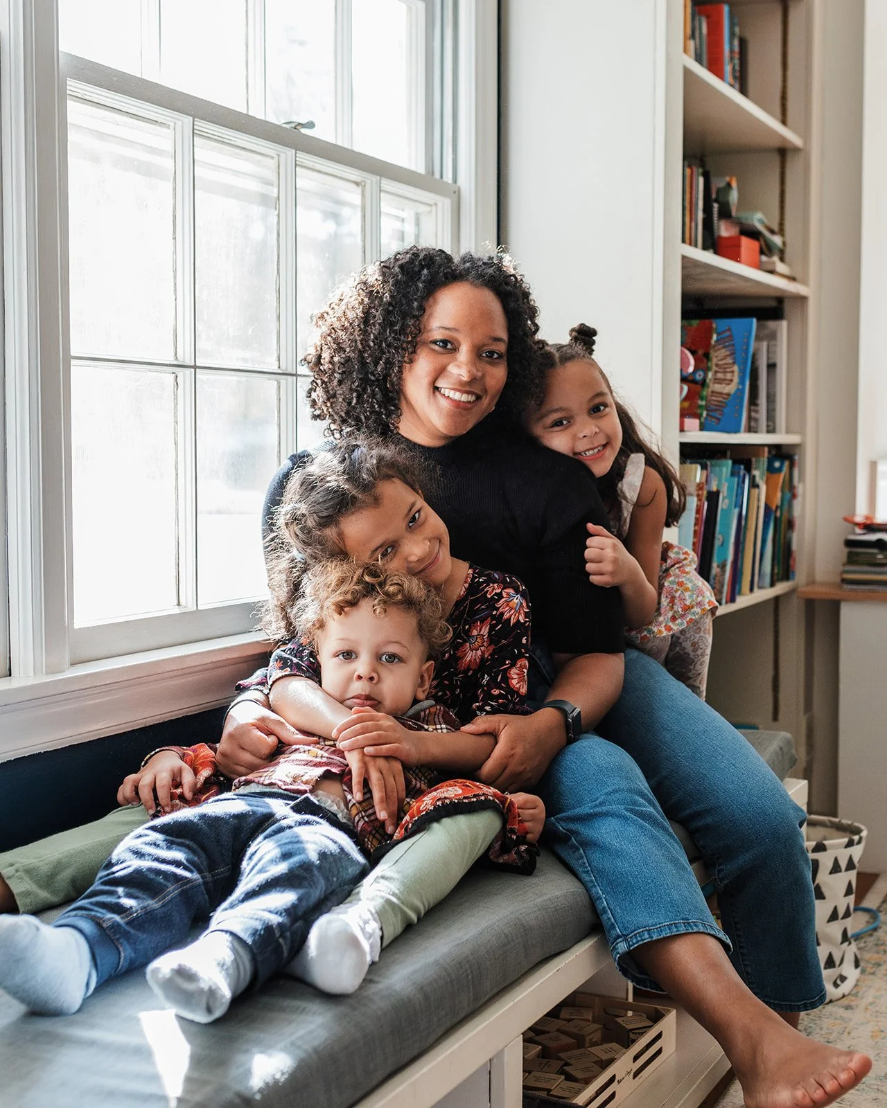 A smiling woman and four children sitting closely together on a bench in front of a window, with bookshelves in the background.