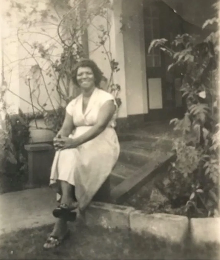 A woman sitting on a low wall outside near plants and trees, smiling at the camera in a vintage black and white photo.