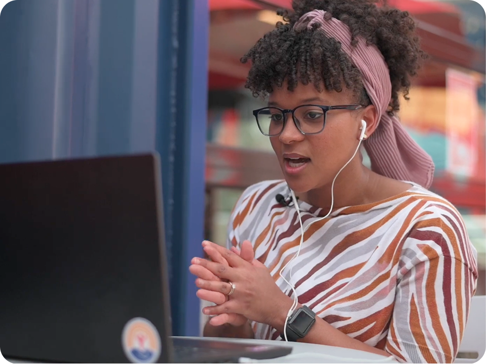 A woman with curly hair, wearing glasses, a striped headscarf, and a patterned shirt, is looking at her laptop screen while wearing headphones.