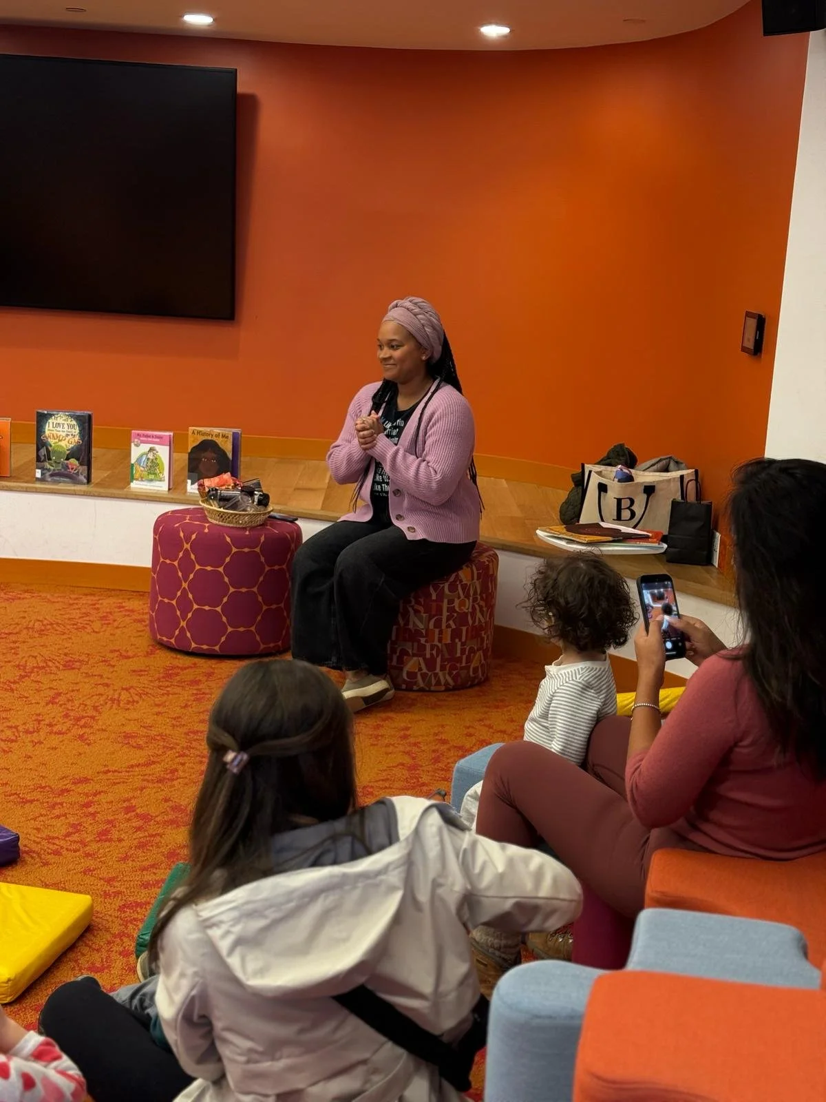 A woman with a pink headwrap and pink cardigan sits on a decorated stool during a storytelling or reading event. She is smiling and engaged with a small audience, including children and adults. Books are displayed on a low shelf, and one audience mem
