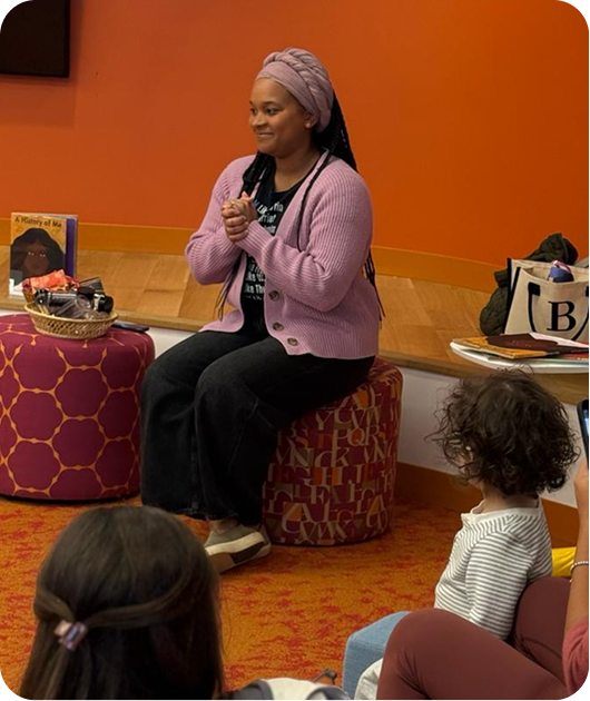 A woman wearing a purple headwrap and matching cardigan sits on a patterned ottoman, speaking to children in a library or bookstore setting.