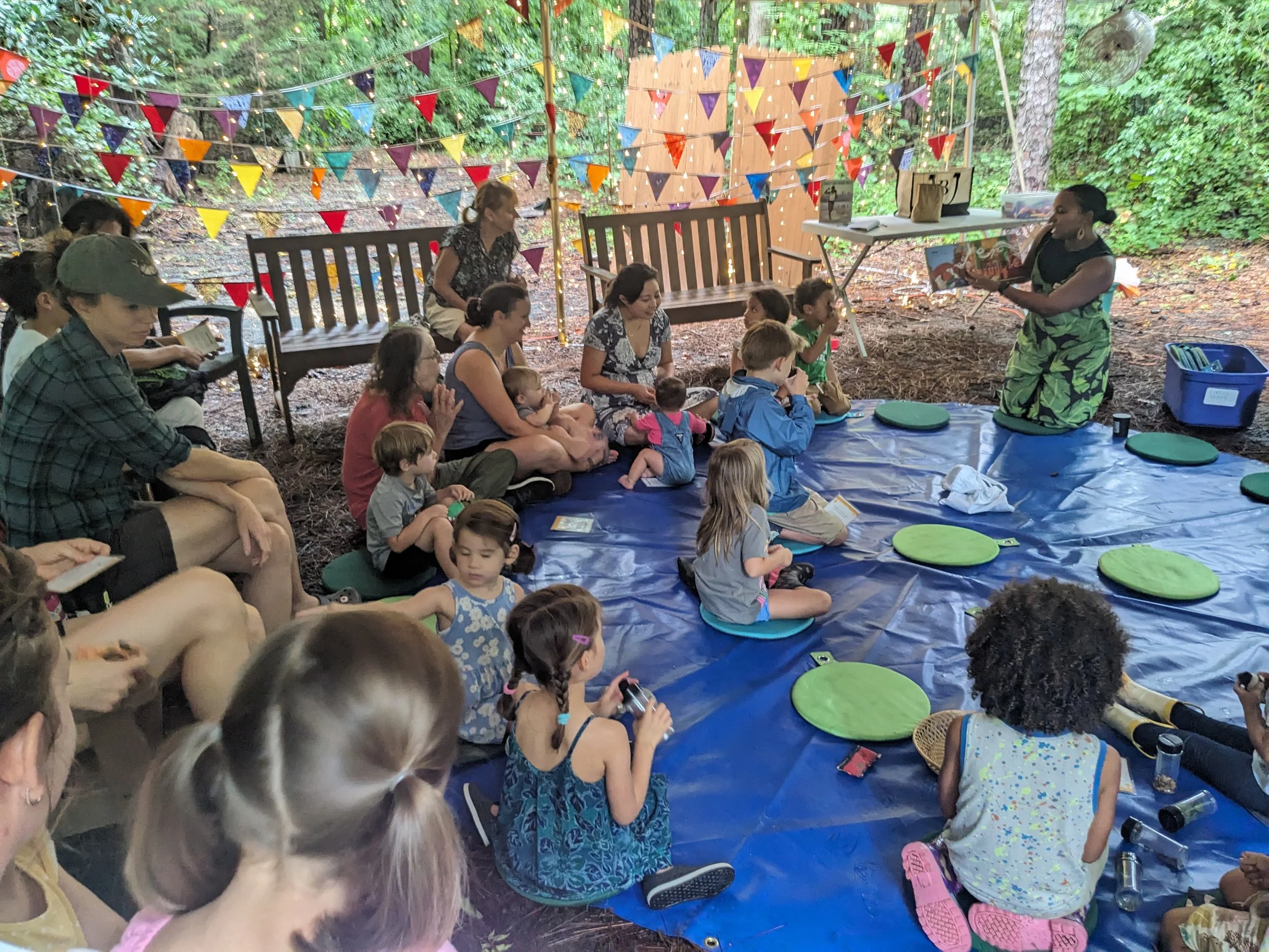 Children and adults sitting on a blue tarp on the ground outdoors, listening to a woman in camouflage pants reading a book. The area is decorated with colorful triangular pennant banners and string lights, and surrounded by trees.