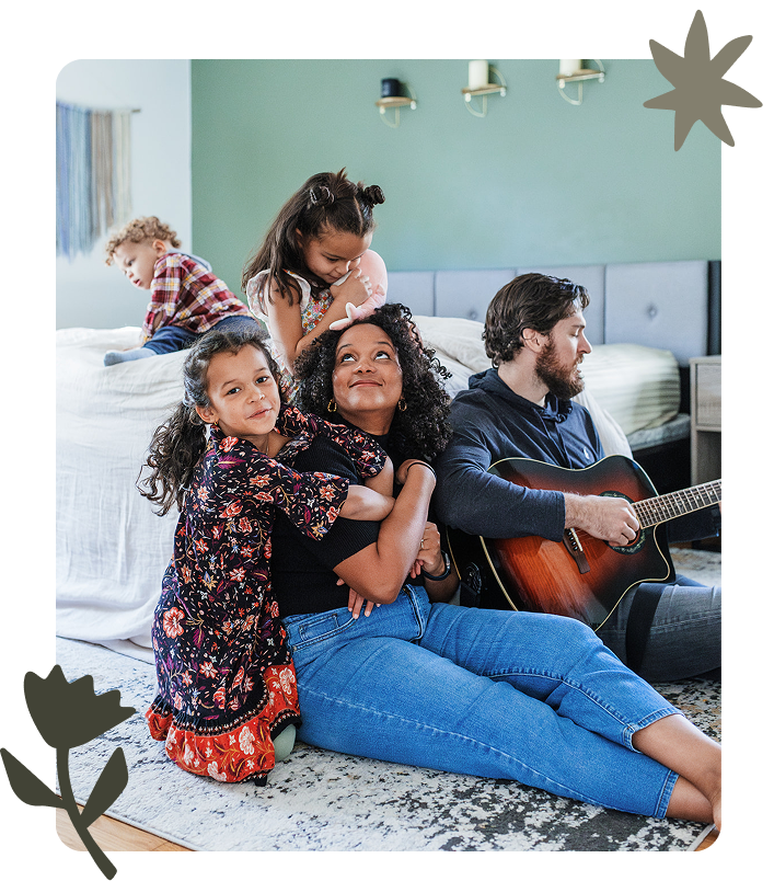 Family spending time together in a cozy living room. A woman is sitting on the floor with two young girls, one hugging her and the other leaning on her shoulder, all smiling and relaxed. In the background, a man is playing guitar, while a boy is sitting on the bed.