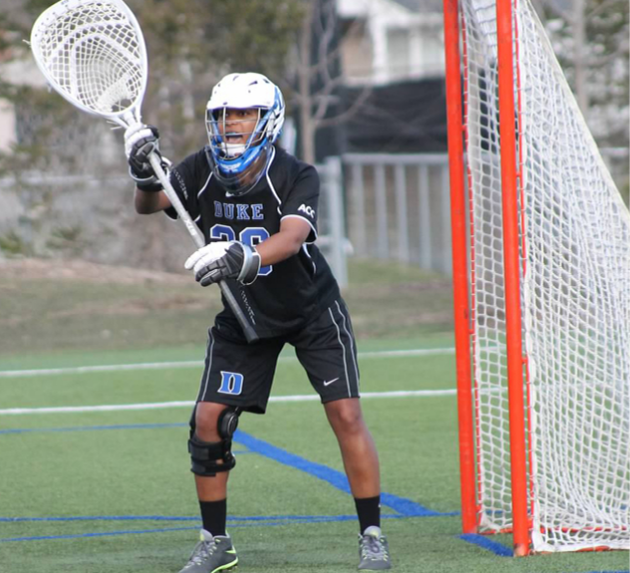 Young lacrosse goalie in black uniform with number 30, wearing a helmet, gloves, and knee pads, holding a lacrosse stick near the goal on a field.