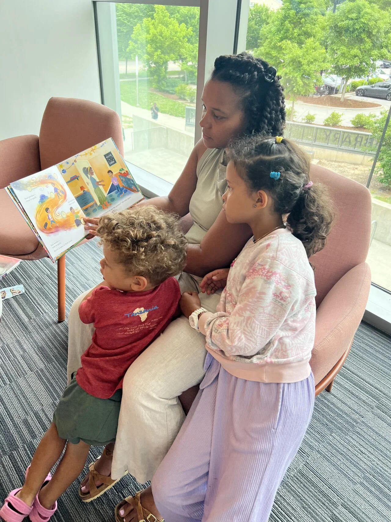 A woman sitting on a pink sofa, reading a children's book with colorful illustrations to two young girls and a boy. One girl has curly hair with colorful clips, and the boy has curly hair and is wearing a red shirt. They are sitting by a large window
