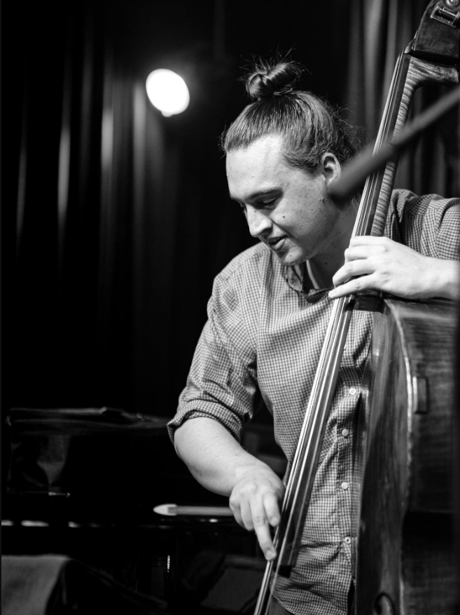 Black and white image of a man with his hair up in a bun plucking the strings on an upright bass