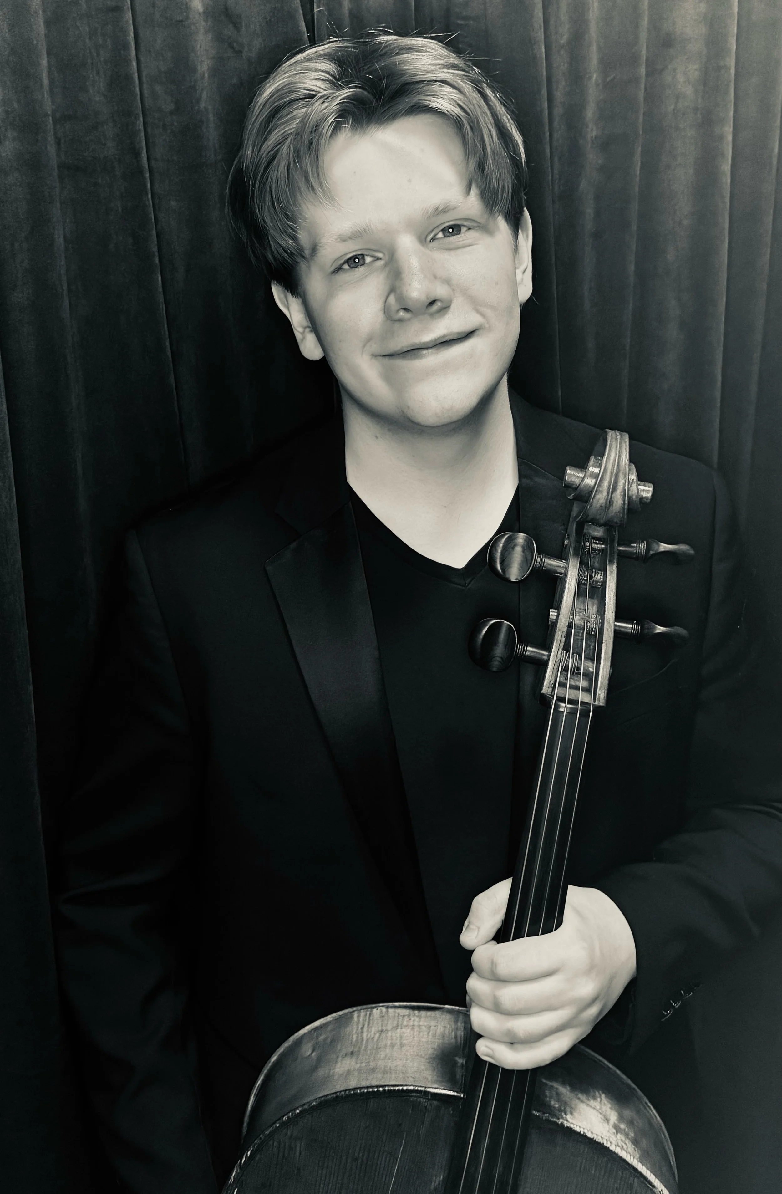 Person holding a cello indoors, smiling, wearing a black shirt.