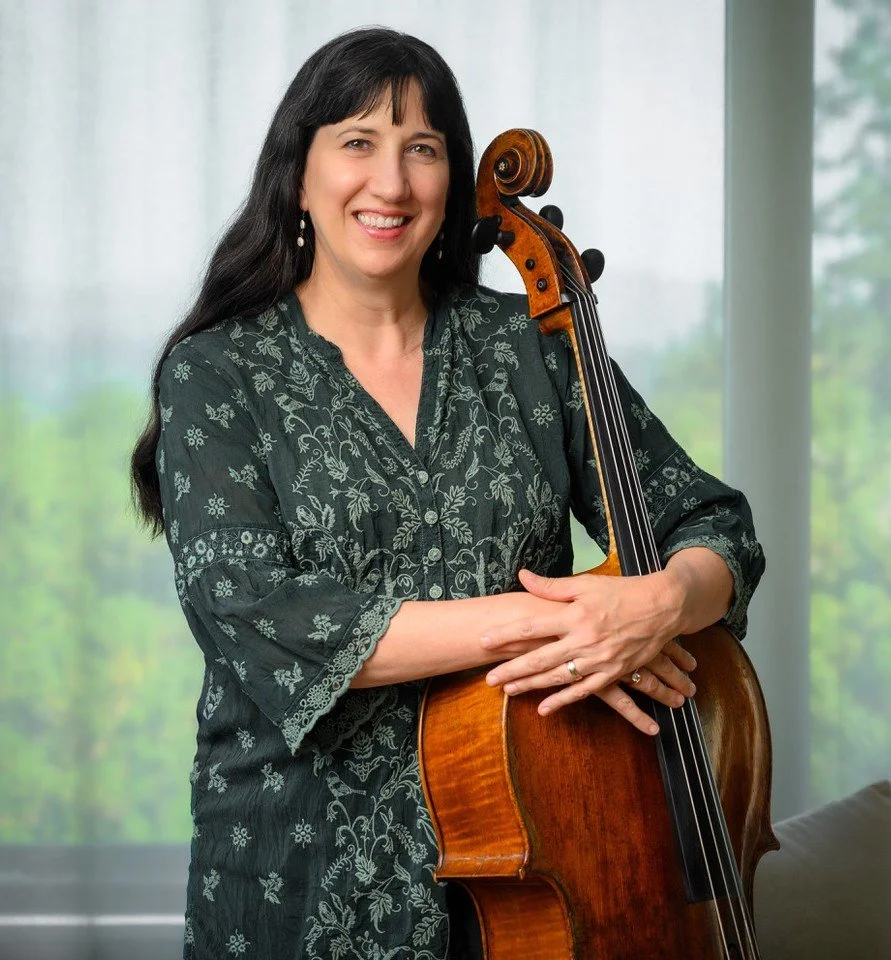 Woman smiling holding a cello in front of a scenic window